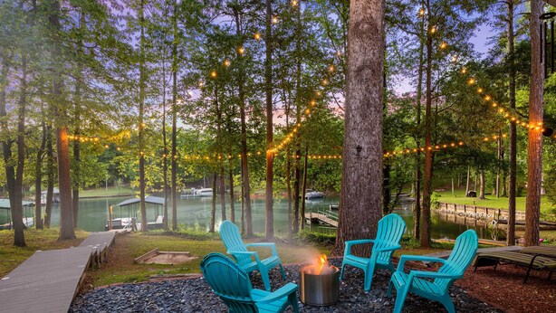 Un espace extérieur aménagé avec des chaises Adirondack, un foyer et des guirlandes lumineuses, le tout dans un cadre boisé.