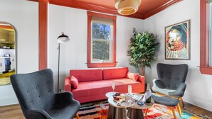 Una sala de estar moderna con un sofá rojo, dos sillones grises, una mesa de centro de madera con bocadillos y bebidas, una lámpara de pie y un ventanal con vistas a árboles.