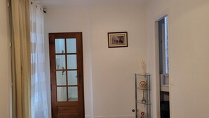 Un couloir avec une porte en bois, un tableau encadré au mur et une étagère en verre ornée d’objets décoratifs.