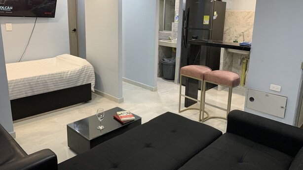 Una habitación de hotel moderna con una cama, un sofá negro, una pequeña mesa de centro con un libro y un vaso, y una zona de cocina con refrigerador y lavaplatos.