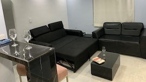 Una sala de estar moderna con un sofá negro, una mesa de centro negra y un mueble para televisor negro. Hay una ventana con cortinas blancas y un techo con iluminación empotrada.