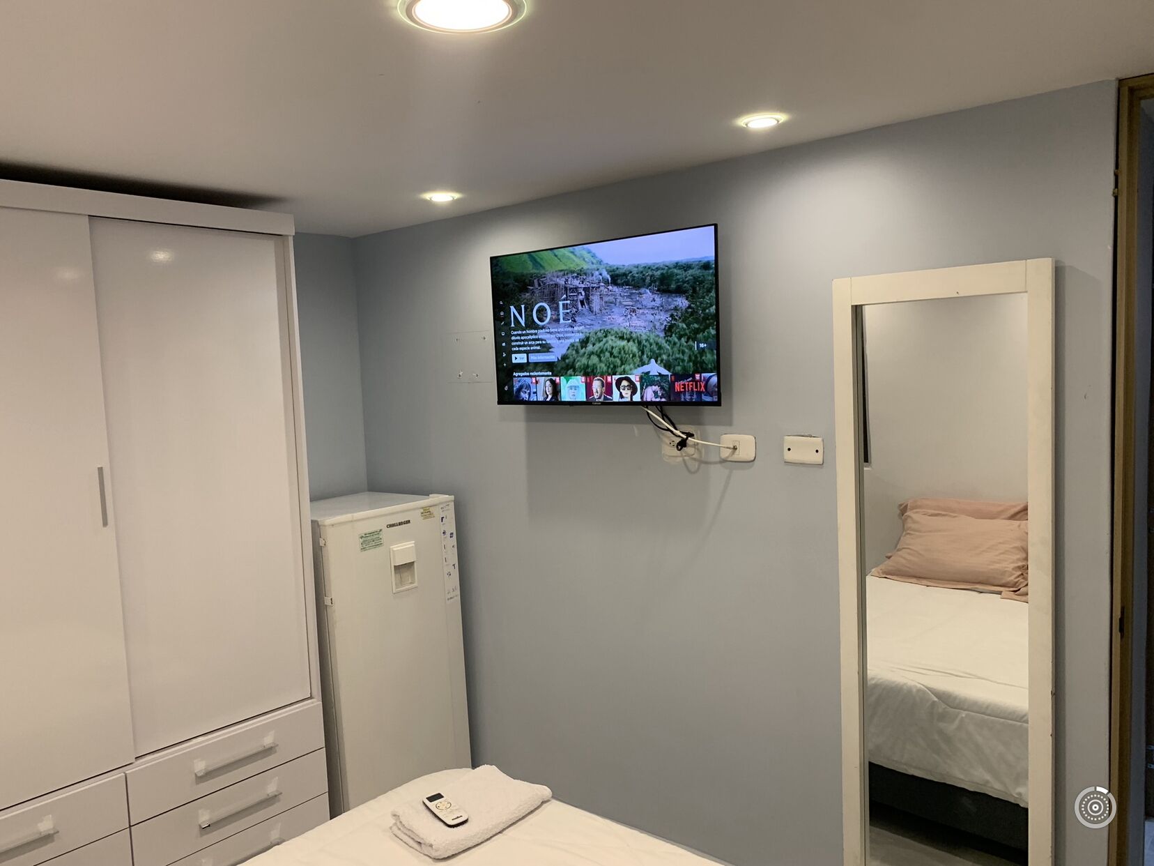 A bedroom with a bed, a TV mounted on the wall displaying a Netflix interface, a bedside table with a lamp, and a wardrobe.