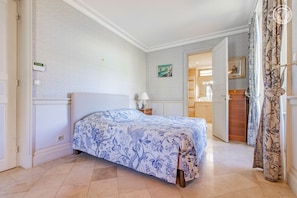 Un dormitorio con cama, mesita de noche, lámpara, un cuadro y un baño visible a través de una puerta abierta.