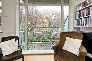 A room with a large window, two armchairs, a bookshelf, and a view of a building and trees outside. - The Little Library (London)