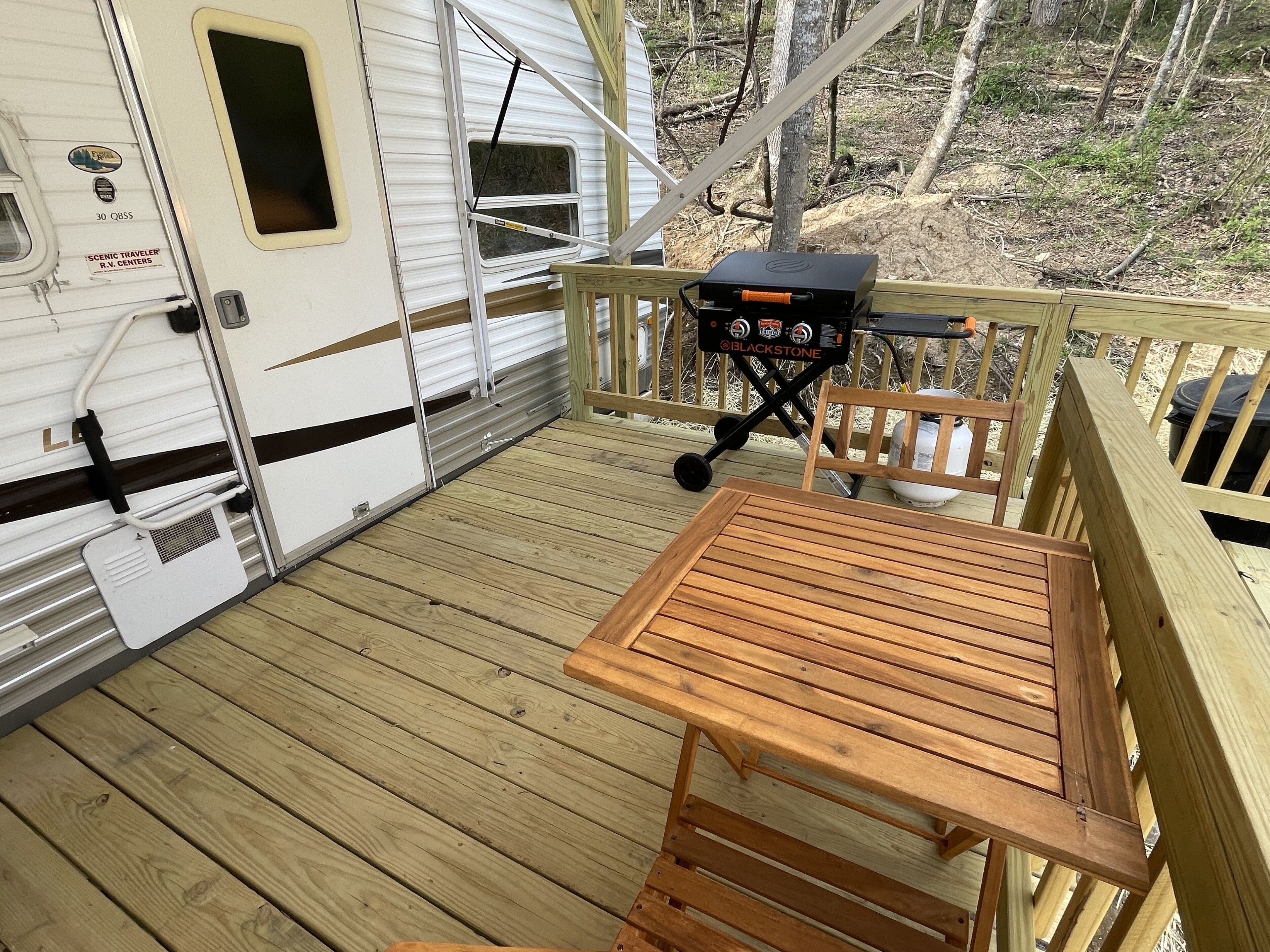 Una terraza de madera con una parrilla y una mesa, junto a una caravana blanca con ventana y puerta.