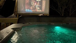 Un jacuzzi con una pantalla grande mostrando una escena de película, rodeado de árboles y luces colgantes.