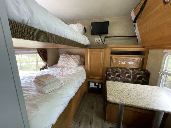 Interior compacto de una caravana con una litera, una pequeña cocina, un televisor y una mesa con una silla.