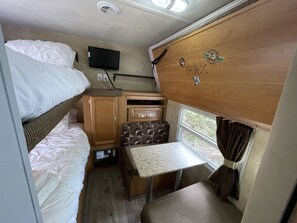 A compact RV interior with bunk beds, a small kitchenette, a television, and a window with curtains. - Ramblin Rose Mountain Camping with Theater, Hot-Tub, Fire-Pit & Hiking Trails (Jonesville)