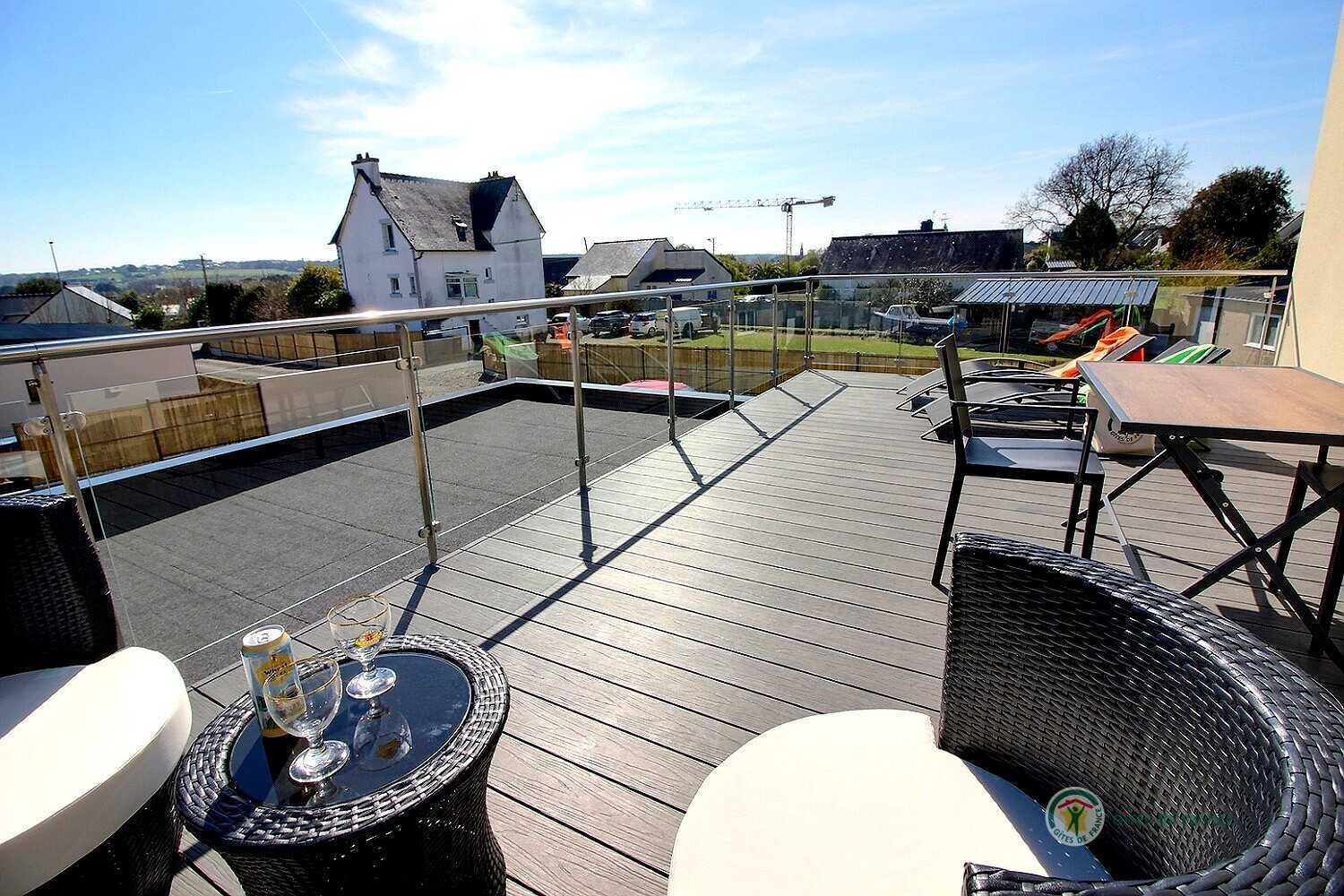 En tagterrasse med udendørs møbler, et bord med drikkevarer og udsigt over det omkringliggende område, inklusive et hus og en byggeplads.