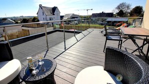 Une terrasse sur le toit, aménagée avec du mobilier d’extérieur, une table avec des boissons, et une vue sur les environs, notamment une maison et un chantier de construction.