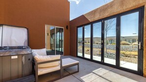 A modern outdoor patio with a hot tub, a cushioned seating area, and large sliding glass doors leading to a patio with a gravel area and buildings in the background.