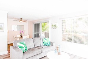 Ein modernes Wohnzimmer mit einem grauen Sofa, einem Deckenventilator, einer Essecke mit Tisch und Stühlen sowie einem großen Fenster mit Jalousien.