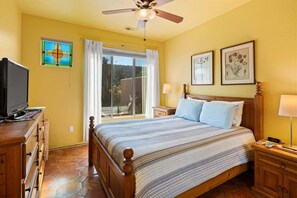 A bedroom with a bed, a television, a ceiling fan, a window with curtains, and framed botanical prints on the wall.