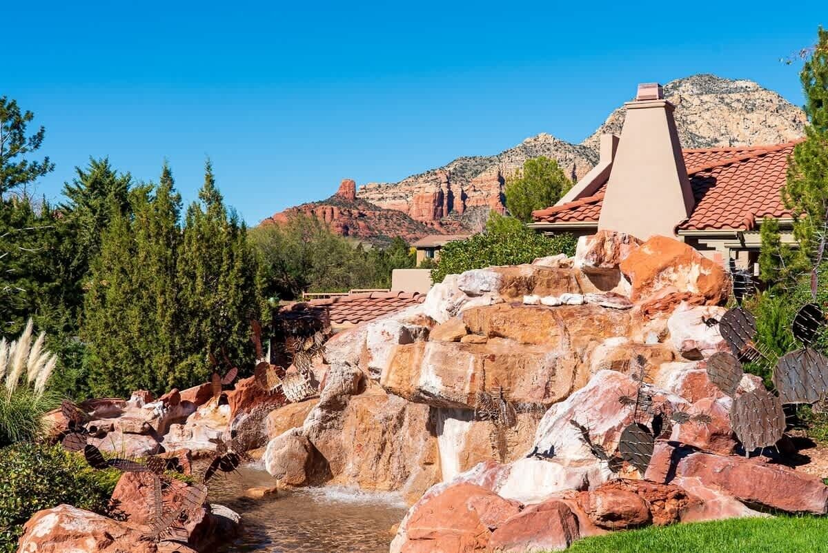 A rock bridge over a stream with a mountainous backdrop and a building with a red-tiled roof.