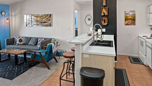 Une cuisine moderne avec un plan de travail blanc, un évier et une cuisinière. Le salon est doté d’un canapé bleu agrémenté de coussins à motifs.