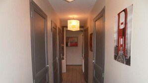 Un couloir étroit au sol carrelé, avec des portes fermées de part et d’autre, et un luminaire suspendu au plafond.