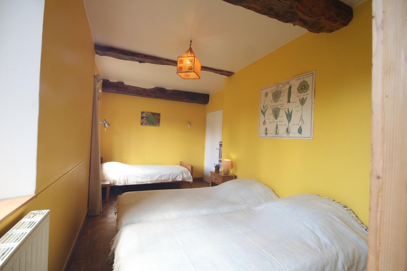 Ein Zimmer mit zwei Betten, einer Holzbalkendecke, einer Wandillustration eines Pflanzenmotivs und einer Hängelampe.