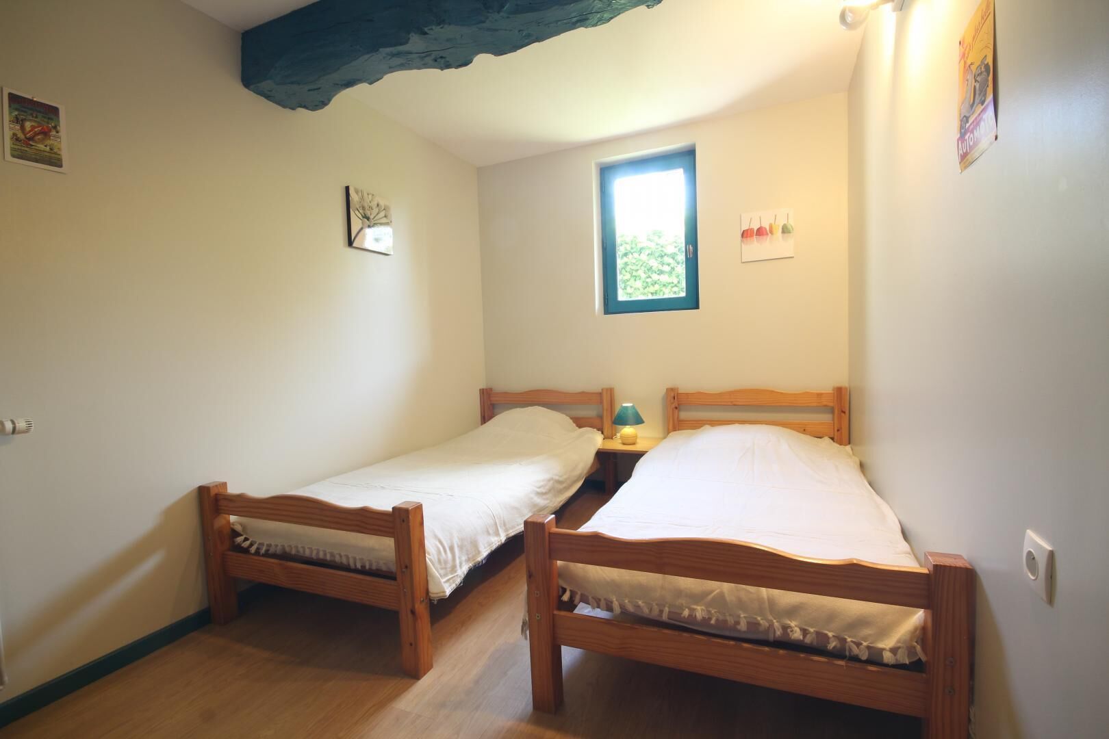 Una habitación pequeña y acogedora con dos camas de madera, una ventana que da a zonas verdes y una mesita con una lámpara.