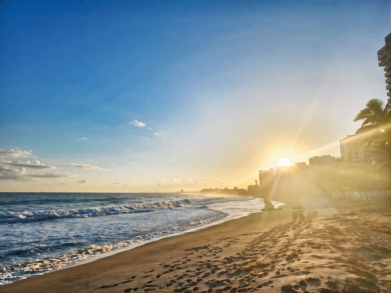 Una playa con huellas que conducen al agua, un atardecer de fondo y un edificio a la derecha.