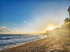 Una playa con huellas que conducen al agua, un atardecer de fondo y un edificio a la derecha.