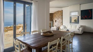 Uma sala de jantar com uma mesa e cadeiras de madeira, vista para o mar através de amplas janelas, uma parede de pedra e um sofá branco ao fundo.