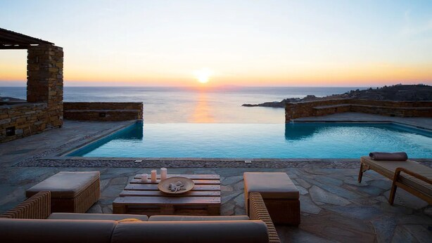 Área de piscina com pilares de pedra, espreguiçadeiras e vista para o pôr do sol sobre o mar.