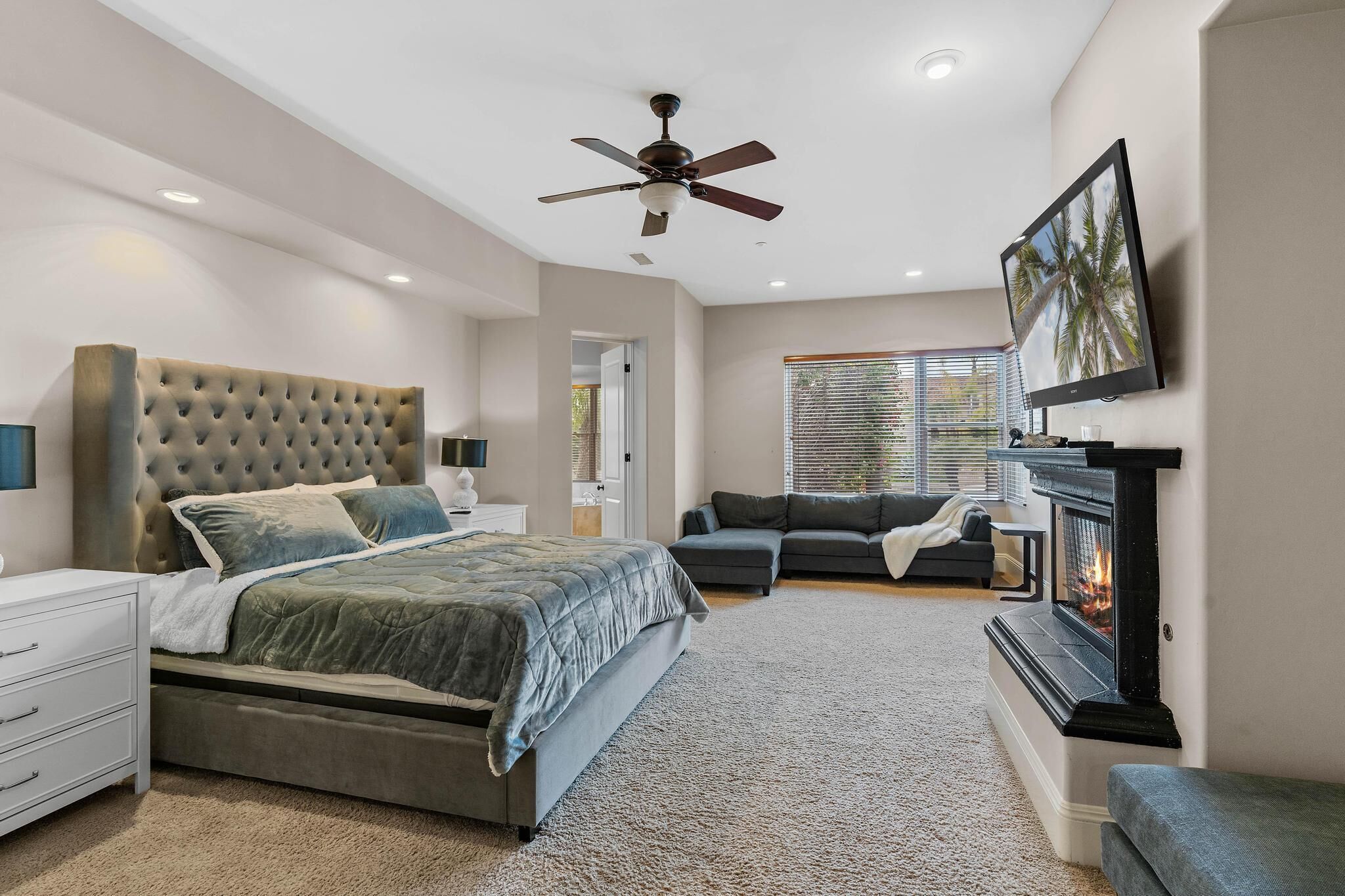 A bedroom with a large bed, a ceiling fan, a fireplace, and a TV on the wall.