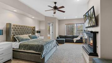 A bedroom with a large bed, a ceiling fan, a fireplace, and a TV on the wall.