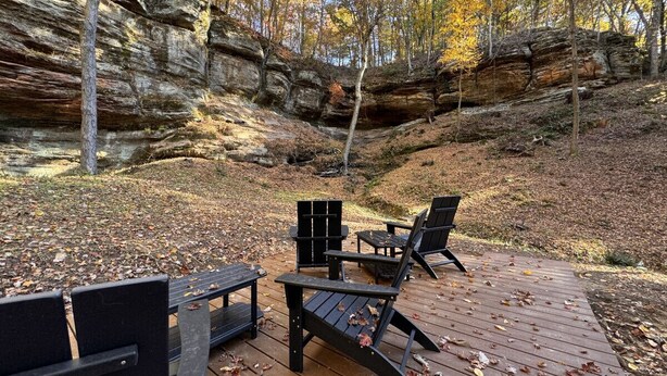 A wooden deck with Adirondack chairs and a table, surrounded by autumn foliage and rocky cliffs. - Modern Cabin w/ Trail to Waterfall/Cave/Cliff (FV) (Rockbridge)