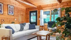 A cozy cabin interior with a wooden floor, a white sofa with blue cushions, a small table, and a large window with a view of greenery. - Modern Cabin w/ Trail to Waterfall/Cave/Cliff (FV) (Rockbridge)