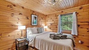 A wooden cabin bedroom with a bed, two bedside lamps, a framed picture on the wall, and a window with curtains. - Modern Cabin w/ Trail to Waterfall/Cave/Cliff (TF) (Rockbridge)