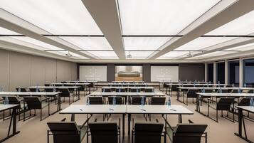 Un salón de conferencias con varias mesas y sillas, una pantalla grande al frente y un proyector.