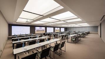 Sala de conferências com várias mesas e cadeiras, amplas janelas e tela de projeção.