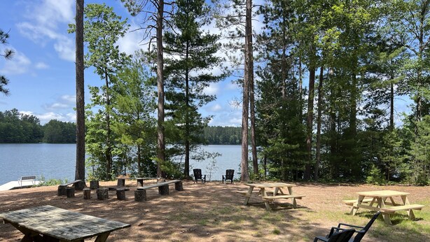 Ein Campingplatz mit Picknicktischen und -stühlen, umgeben von hohen Bäumen und einem See.