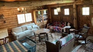 Ein rustikales Blockhaus-Interieur mit einem weißen Sofa, Holzstühlen, einem Esstisch und einer bunten Decke.