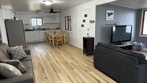 Un salon moderne avec un canapé gris foncé, une table à manger en bois avec des chaises, un coin cuisine avec des meubles blancs et un ventilateur de plafond.
