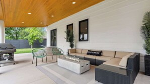 Una terrazza esterna con tetto in legno, un divano con un cuscino con la scritta 'HELLO SUMMER', un tavolino da caffè con piano in vetro e un barbecue.