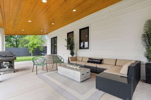 Une terrasse extérieure dotée d’un plafond en bois, d’un canapé agrémenté d’un coussin orné de l’inscription « BONJOUR L’ÉTÉ », d’une table basse avec un plateau en verre et d’un barbecue.