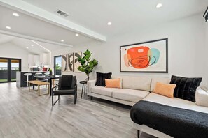 Un salon moderne avec un canapé, un fauteuil et une table basse. On y trouve également un coin repas avec une table et des chaises. Une cuisine est visible au fond.