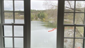 Uitzicht door een ruit met meerdere vlakken, op kalm water met een rood voorwerp dat erop drijft, omringd door bomen en enkele gebouwen.