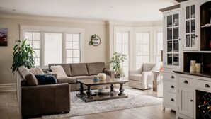 Een ruime woonkamer met een bruine bank, een salontafel en een witte fauteuil. Er is een groot raam met witte gordijnen, een potplant en een kast met glazen deuren.