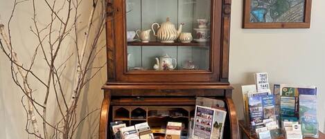 Un mueble de madera con puertas de cristal que exhibe vajilla y teteras, una pequeña mesa con folletos y un cuadro decorativo en la pared.