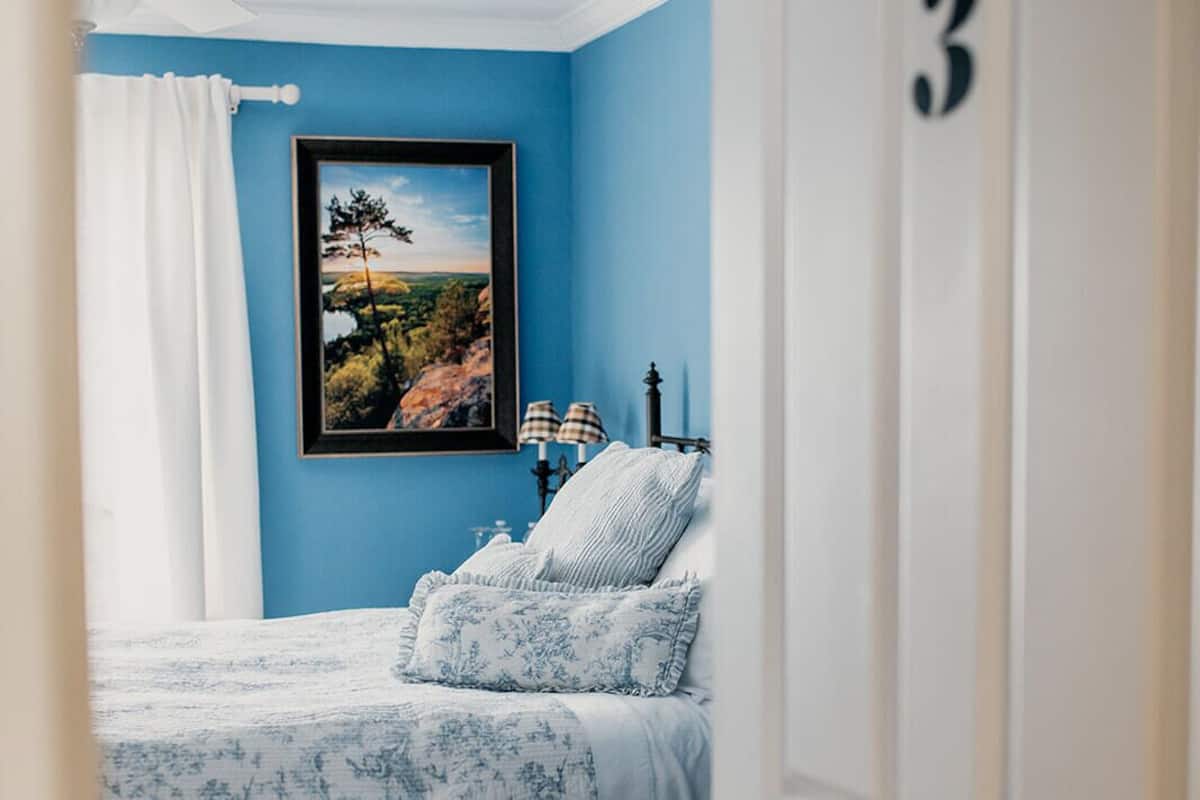 A bedroom with a blue wall, a bed with floral bedding, a framed landscape picture, and a door with the number 3.