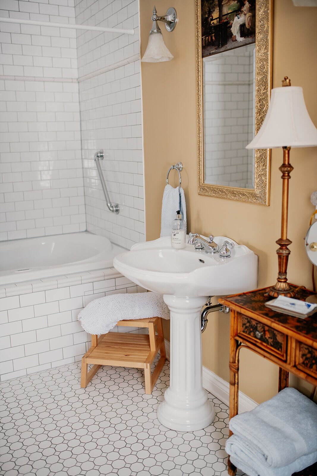 Un baño con una bañera blanca, un lavamanos pedestal, un taburete de madera, un espejo con marco dorado y una lámpara de pared.