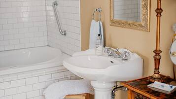 Un baño con una bañera blanca, un lavamanos pedestal, un taburete de madera, un espejo con marco dorado y una lámpara de pared.