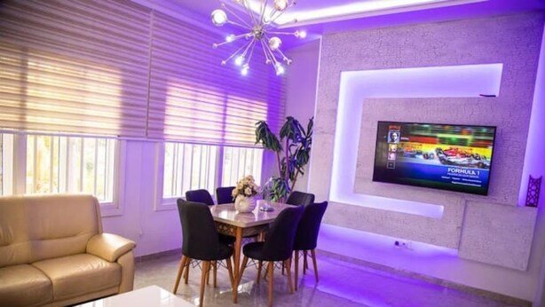 Un salon moderne comprenant un coin repas, un téléviseur à écran plat fixé au mur et une grande fenêtre équipée de stores.
