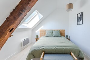 Une chambre avec un plafond en pente, un puits de lumière, un lit avec une tête de lit en bois, deux tables de chevet avec des lampes et un tableau encadré au mur.