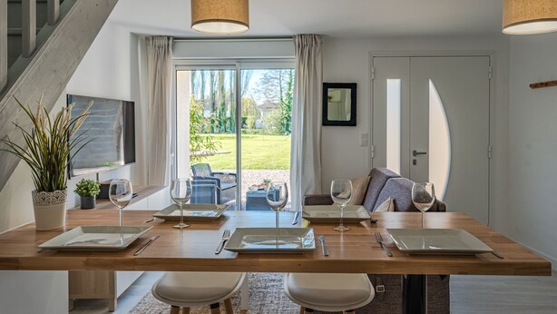 A modern dining area with a wooden table set for four, two chairs on each side, and a view of a garden through large glass doors. - La Grange du Noé: Le Noé (Caen)