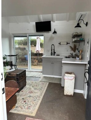 A compact kitchen with a built-in sink, a small dining area with a table and chairs, and a view through a sliding glass door to an outdoor area. - Buckley Farm Casita (Menifee)