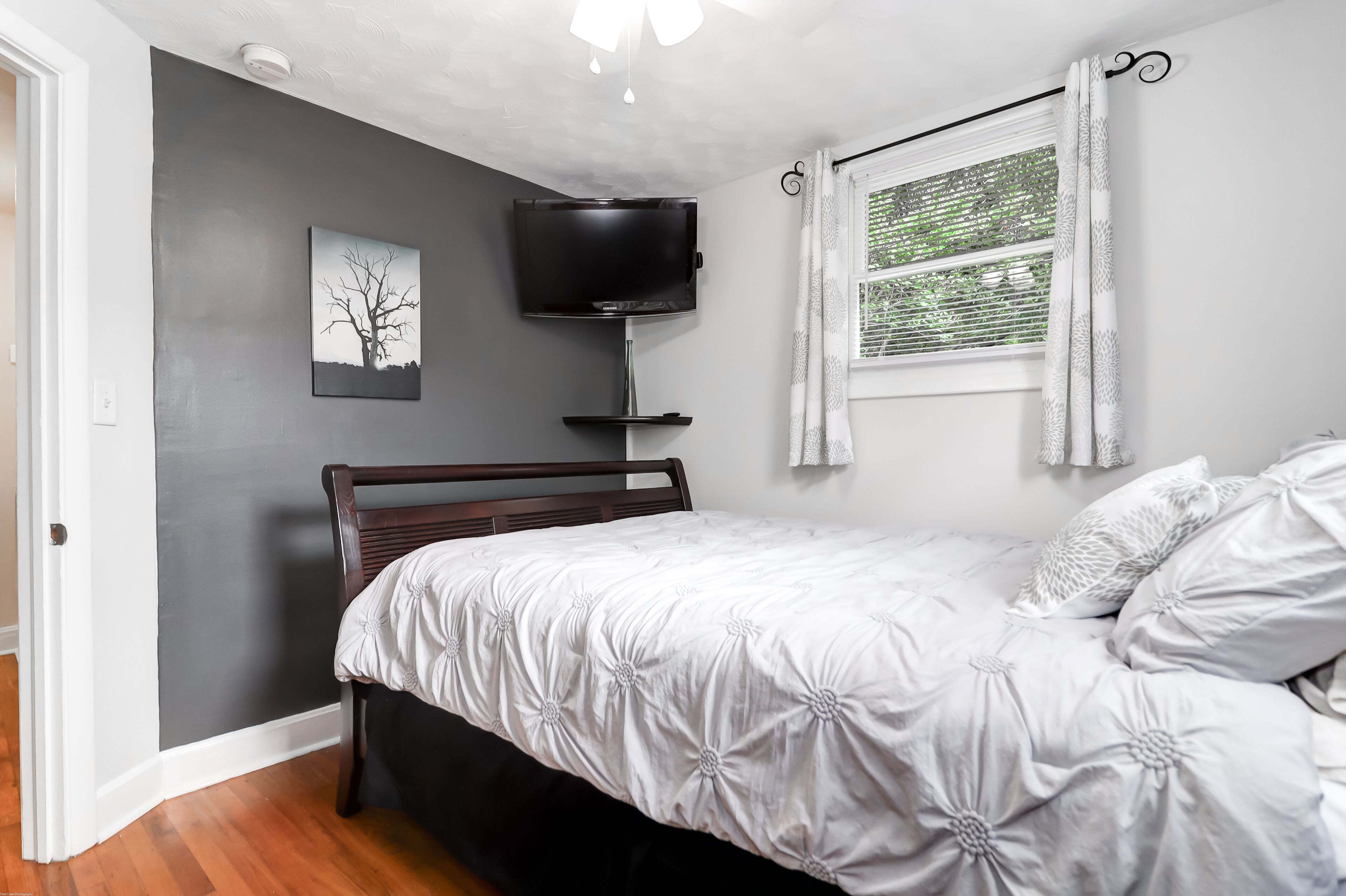 Un dormitorio con cama, televisor montado en la pared, ventana con cortinas e imagen colgada en la pared.